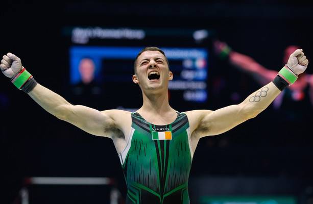 A male gymnast in a green and black leotard with an Irish flag, celebrating with arms outstretched.
