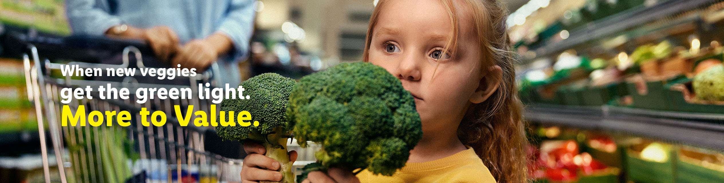 A young girl in a grocery store holding two heads of broccoli, with a shopping cart and adult in the background.