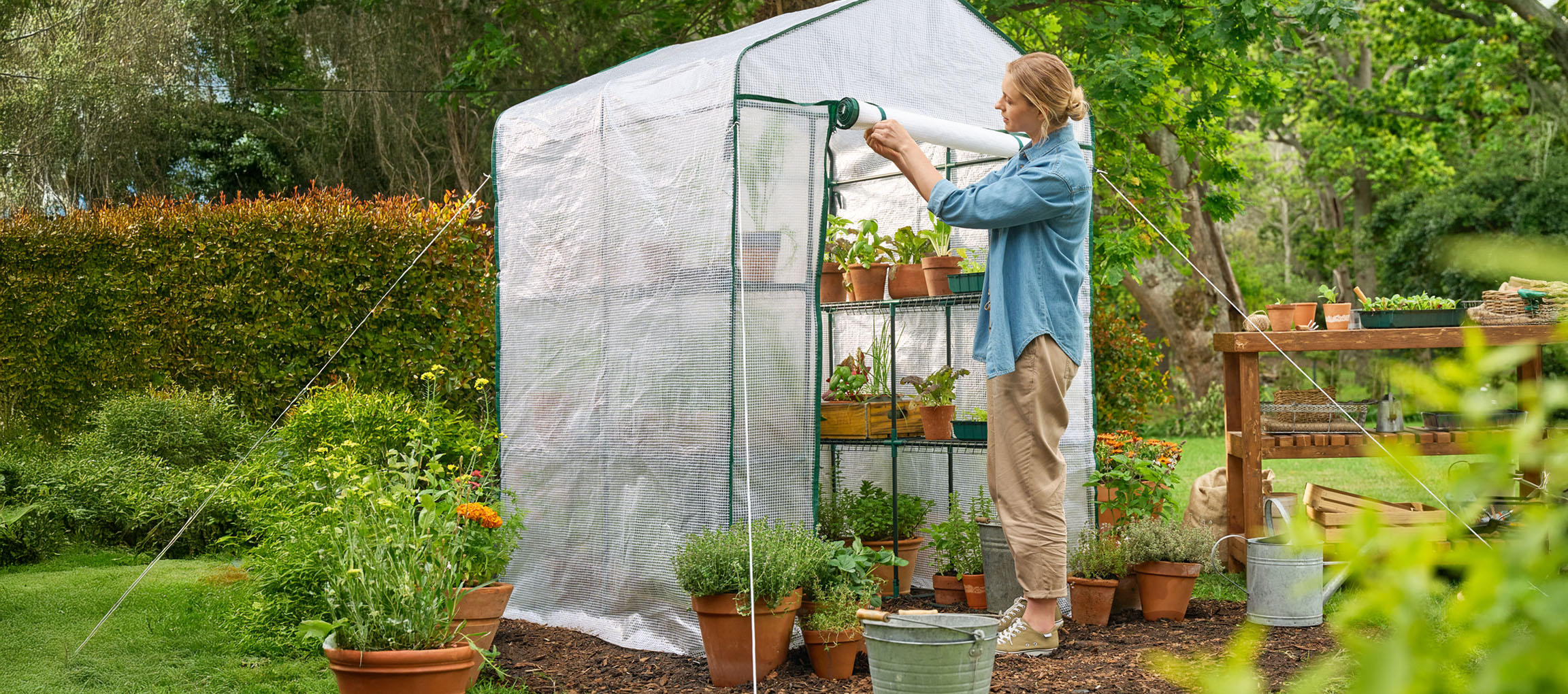 Woman opening a greenhouse with plants and pots in a garden.