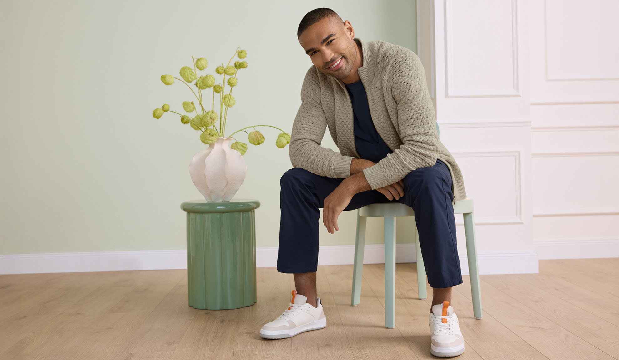Man in a beige cardigan, navy t-shirt, and trousers, sitting on a stool.
