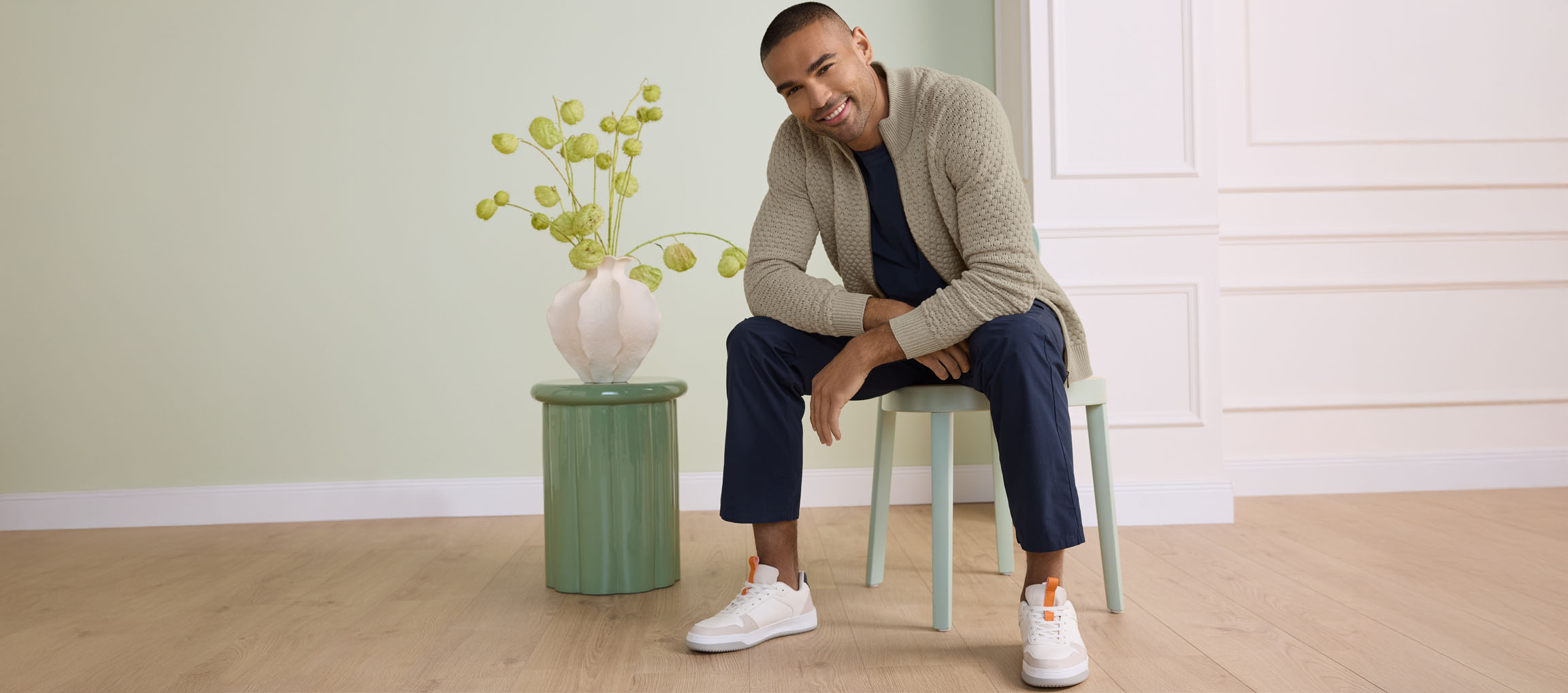 Man sitting on a chair, wearing a cardigan, t-shirt, trousers, and sneakers.