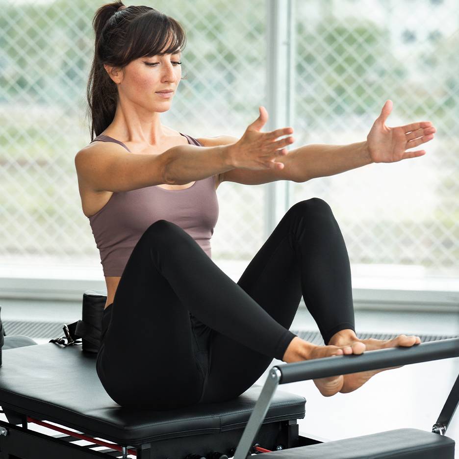 Woman doing Pilates on a reformer, wearing a top and black leggings.