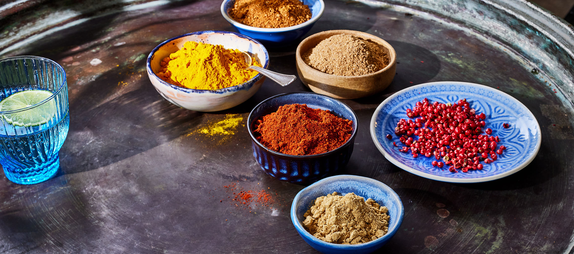 Various spices, including turmeric, paprika, and pink peppercorns, in bowls on a dark surface.