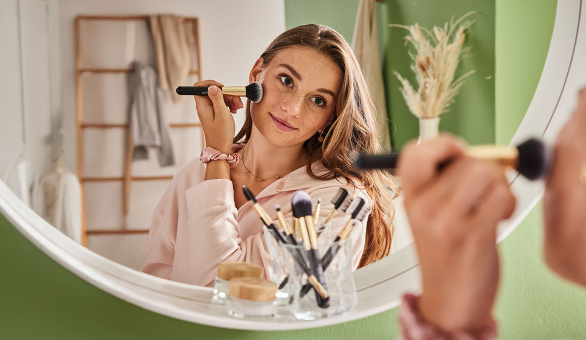 Woman applying makeup with a brush, reflected in an oval mirror.