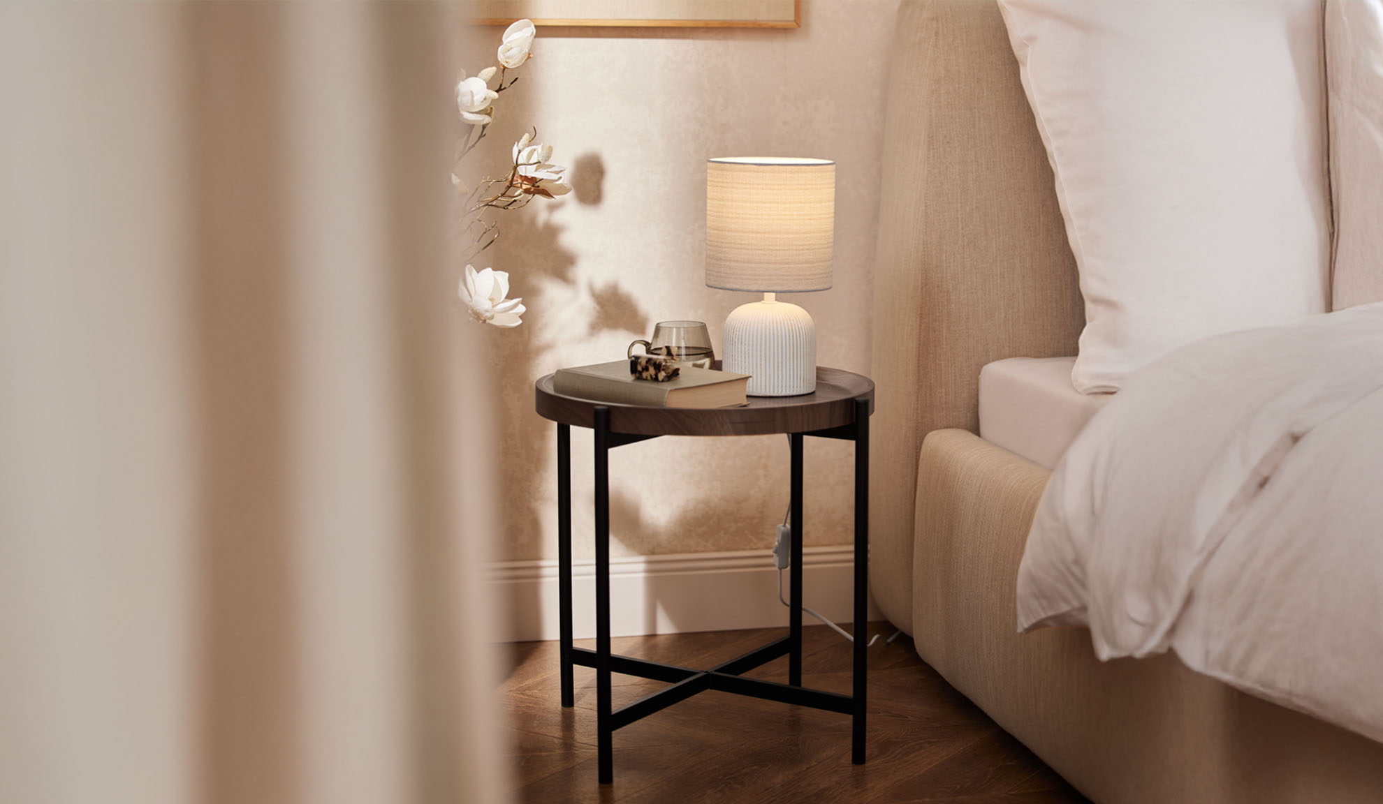 Round wooden nightstand with a table lamp and accessories next to a bed.