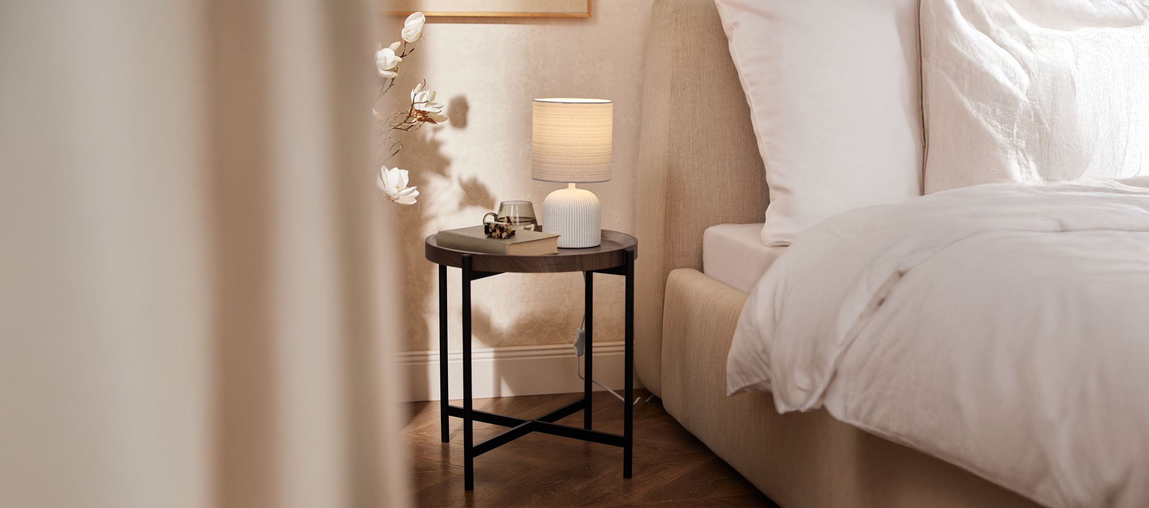 Round wooden nightstand with a table lamp and a beige bed in a cozy bedroom.
