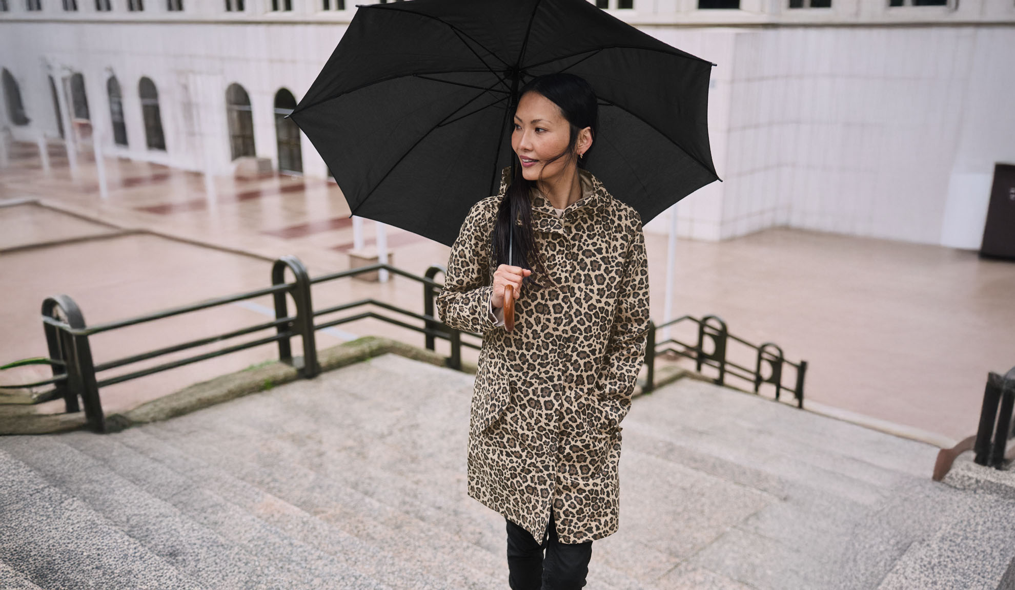 A woman in a leopard print coat holds a black umbrella on a staircase.