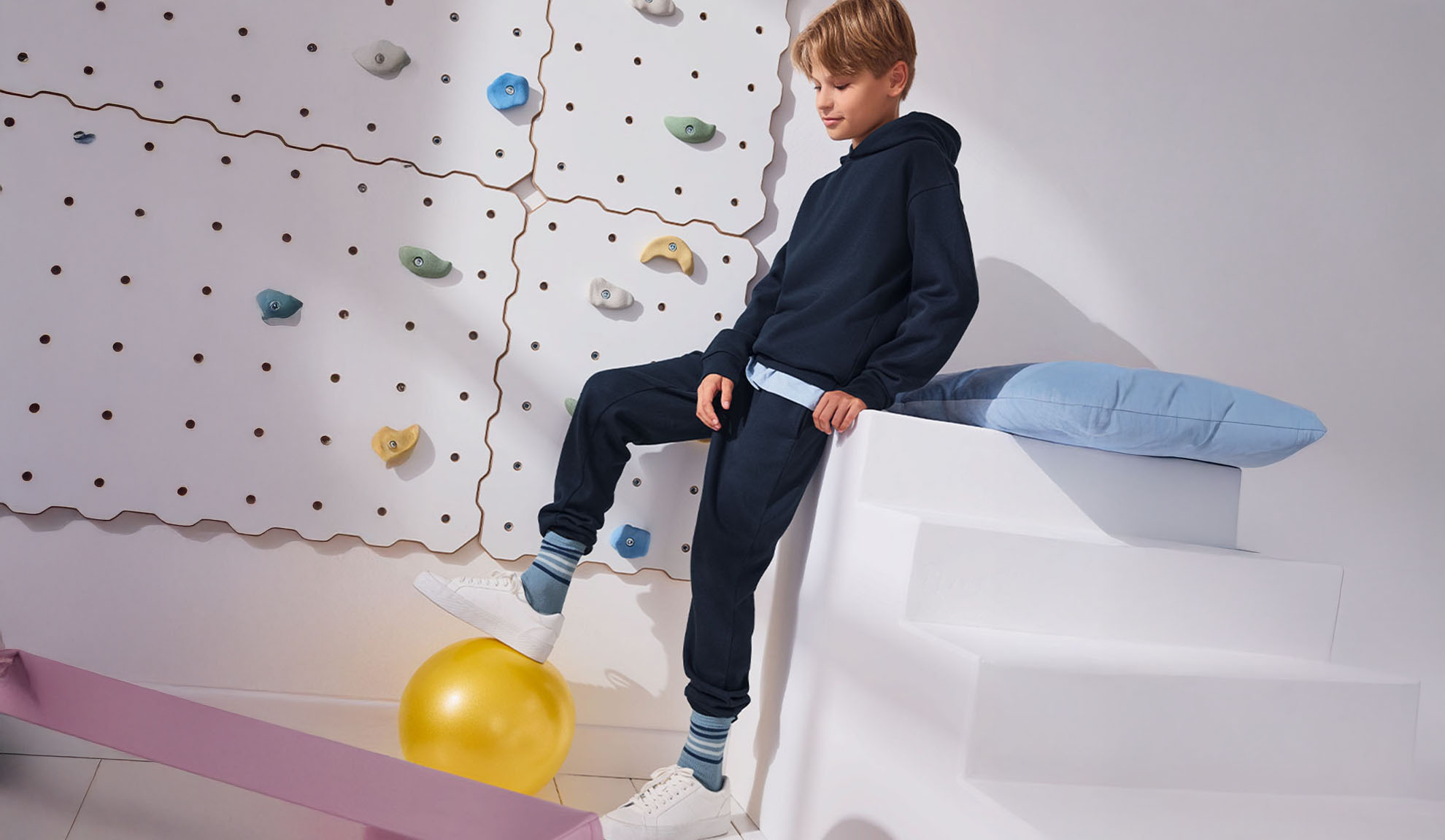 A boy stands on an exercise ball next to a climbing wall, wearing a hoodie and sweatpants.