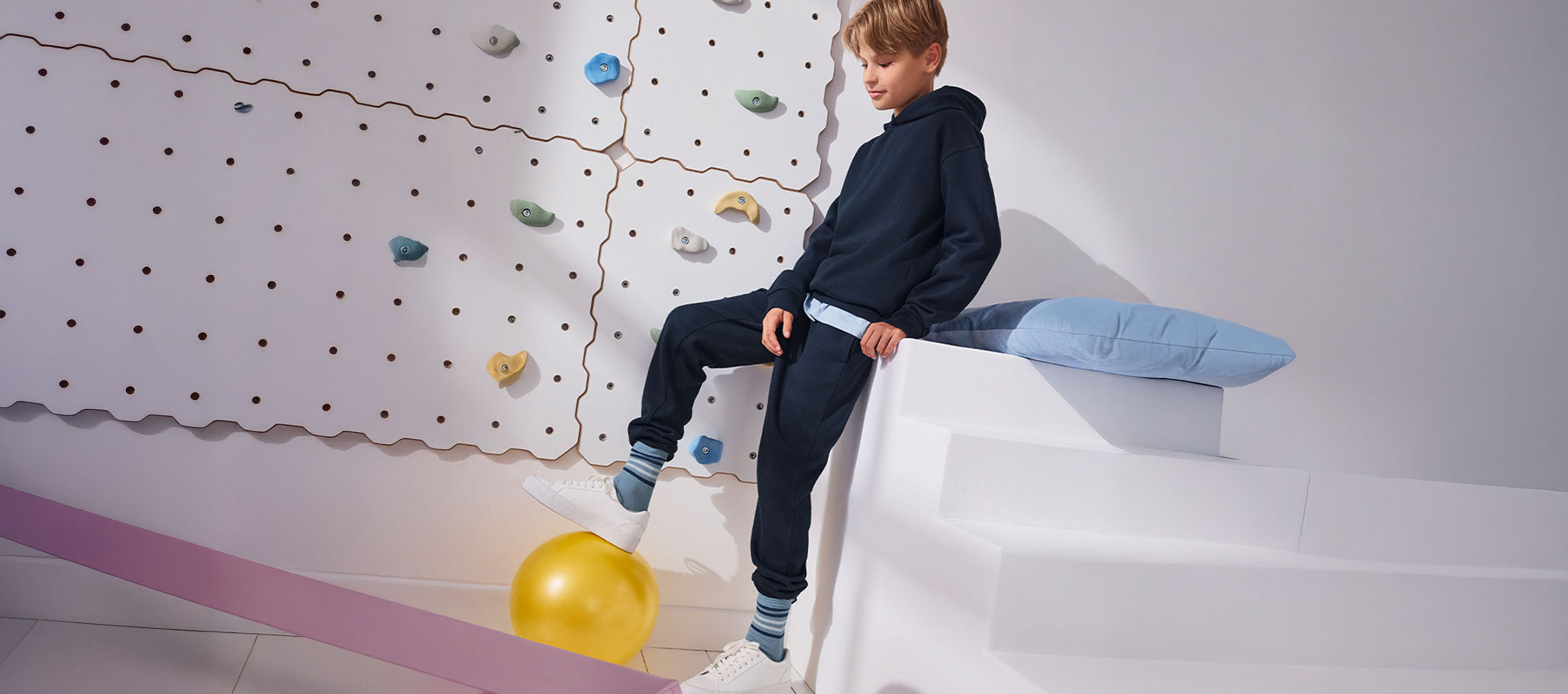 A boy in a navy hoodie and sweatpants, with one foot on a yellow ball, next to a climbing wall.