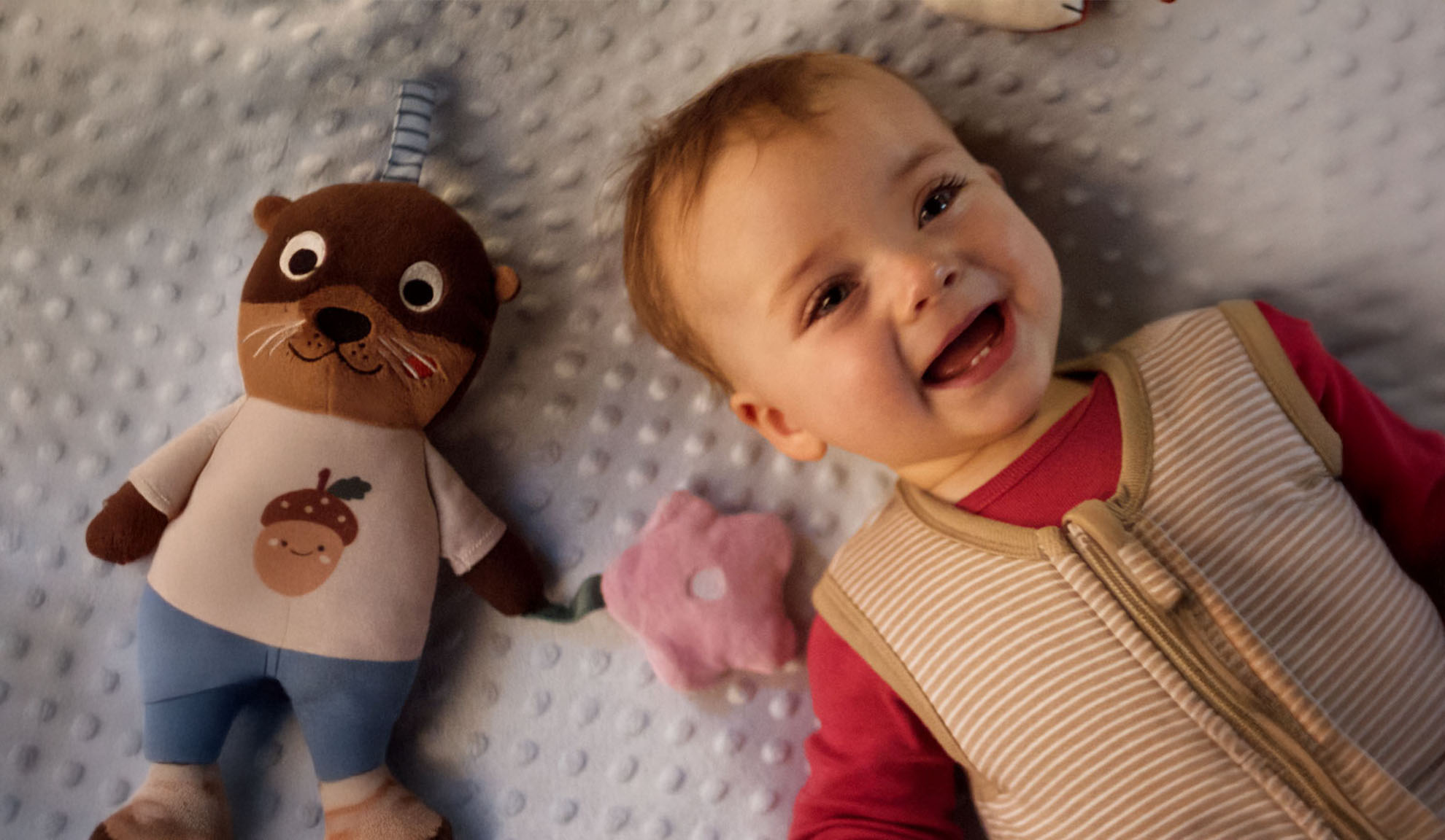 A smiling baby and an otter plush toy on a soft blanket.