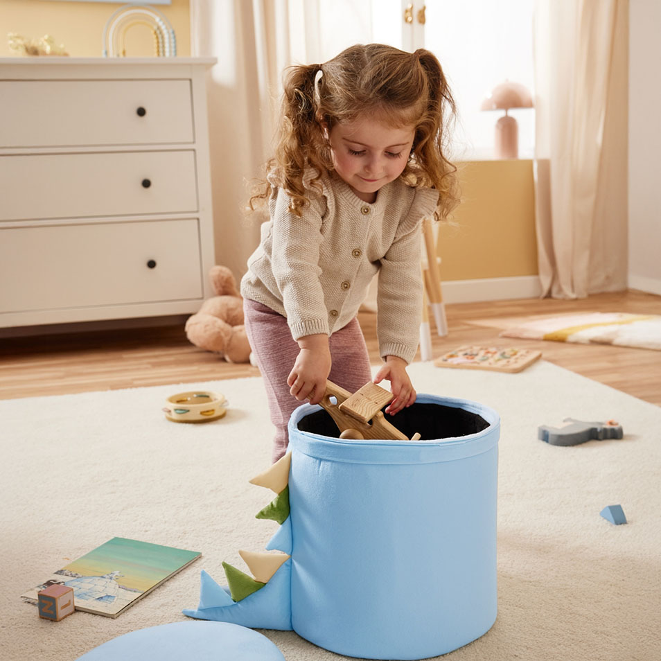 A child puts wooden toys into a blue dinosaur-themed storage bin in a bedroom.