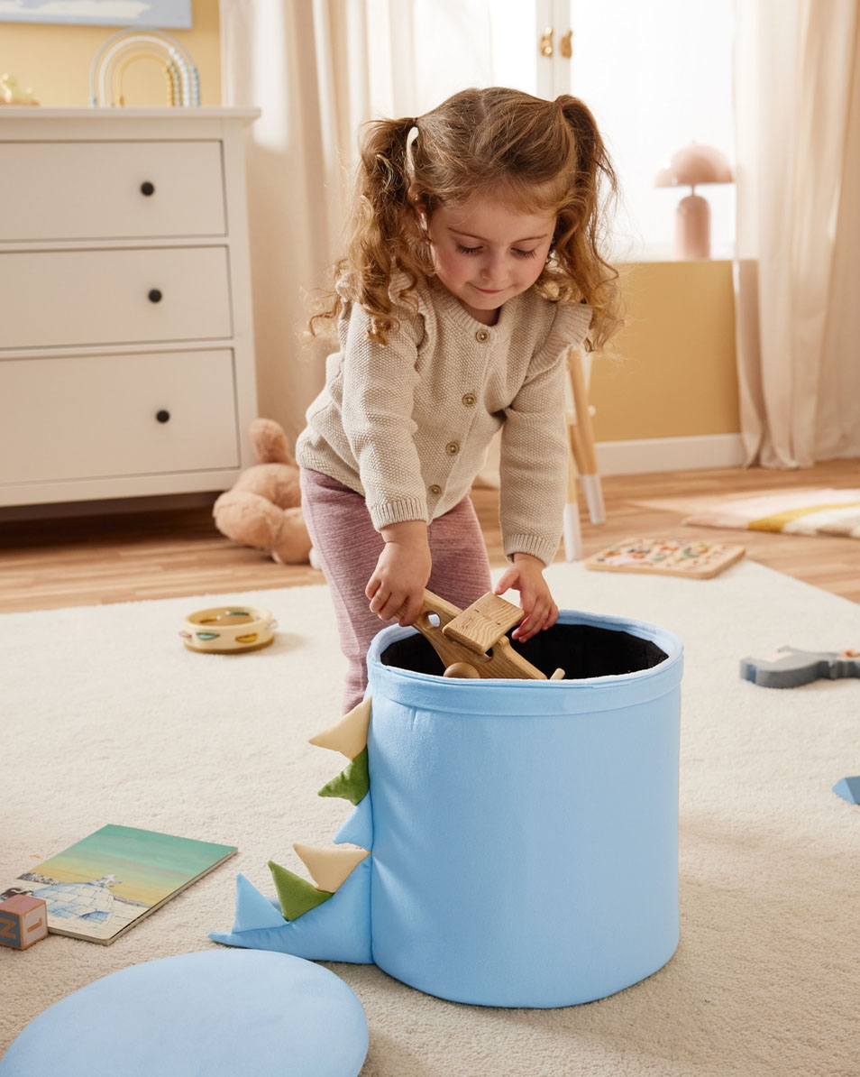 A child puts wooden toys into a blue dinosaur-themed storage bin.