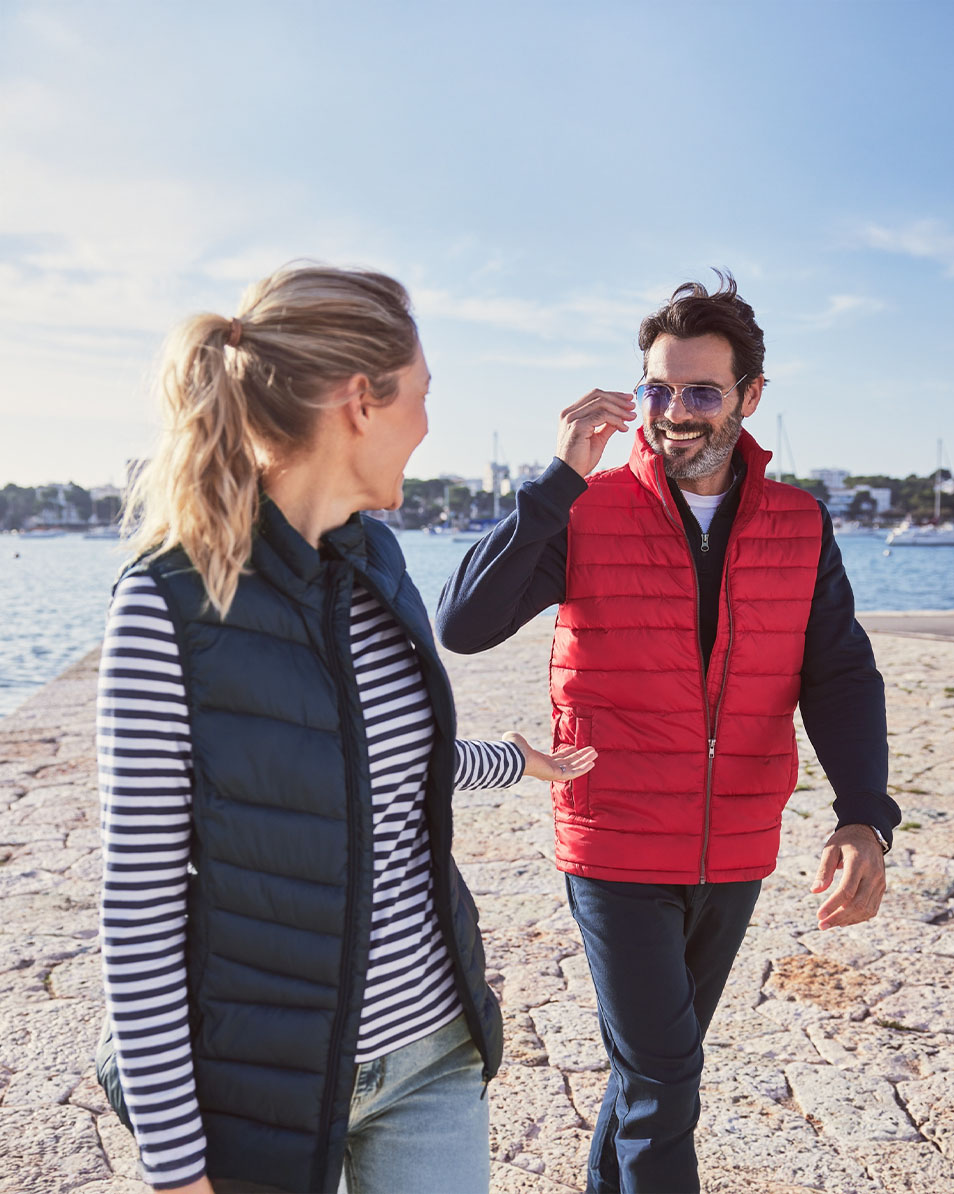 A smiling couple wears quilted vests on a sunny quay.
