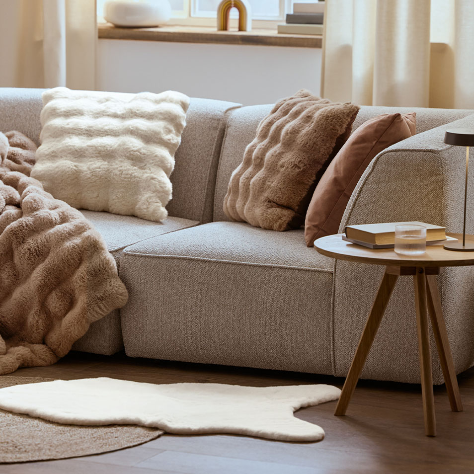 A gray modular sofa with fluffy pillows and a blanket, next to a wooden side table.