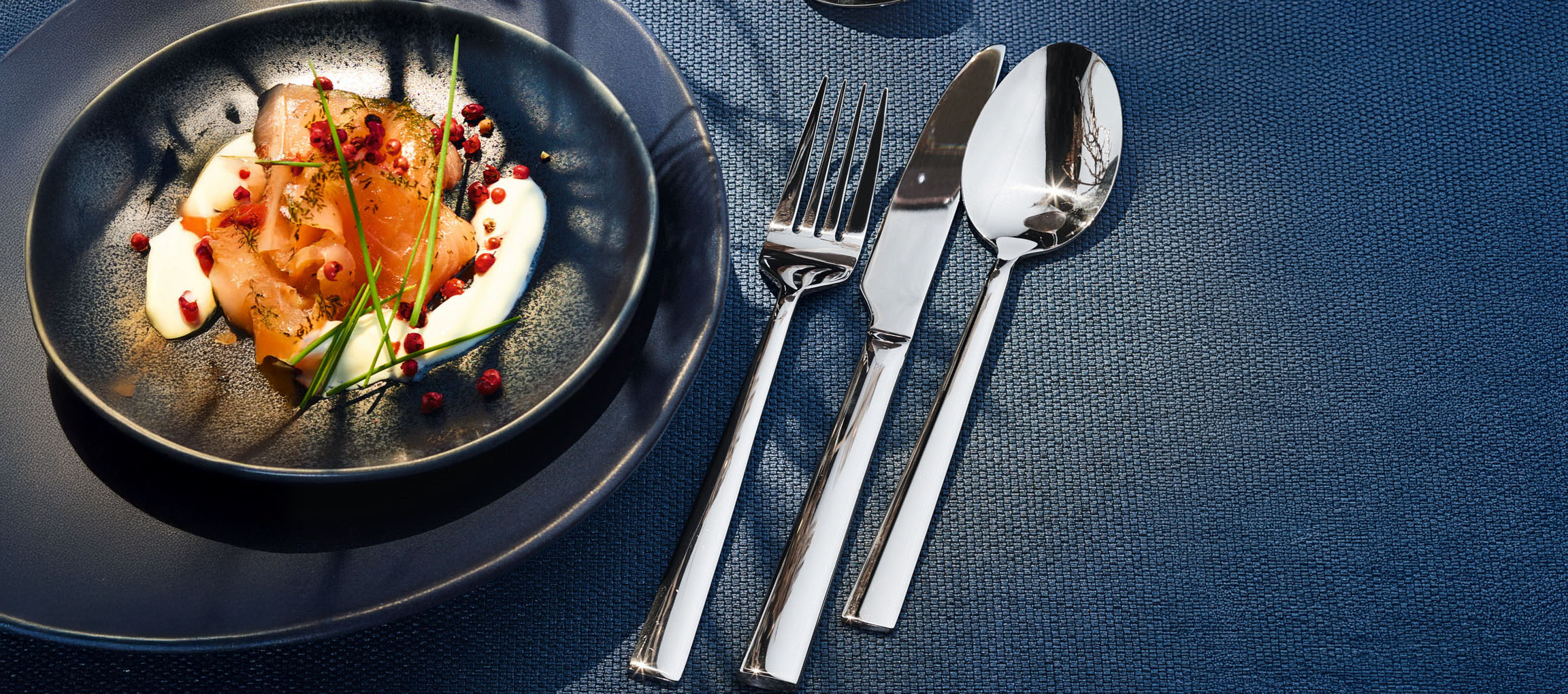 Smoked salmon with cream, chives, and pink peppercorns on a dark plate with cutlery.