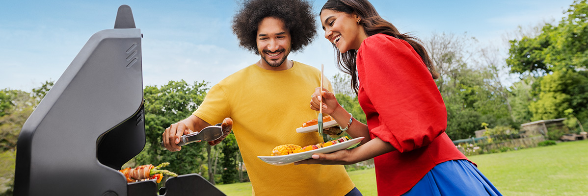 A couple grilling food outdoors, with a man using tongs and a woman holding a plate.