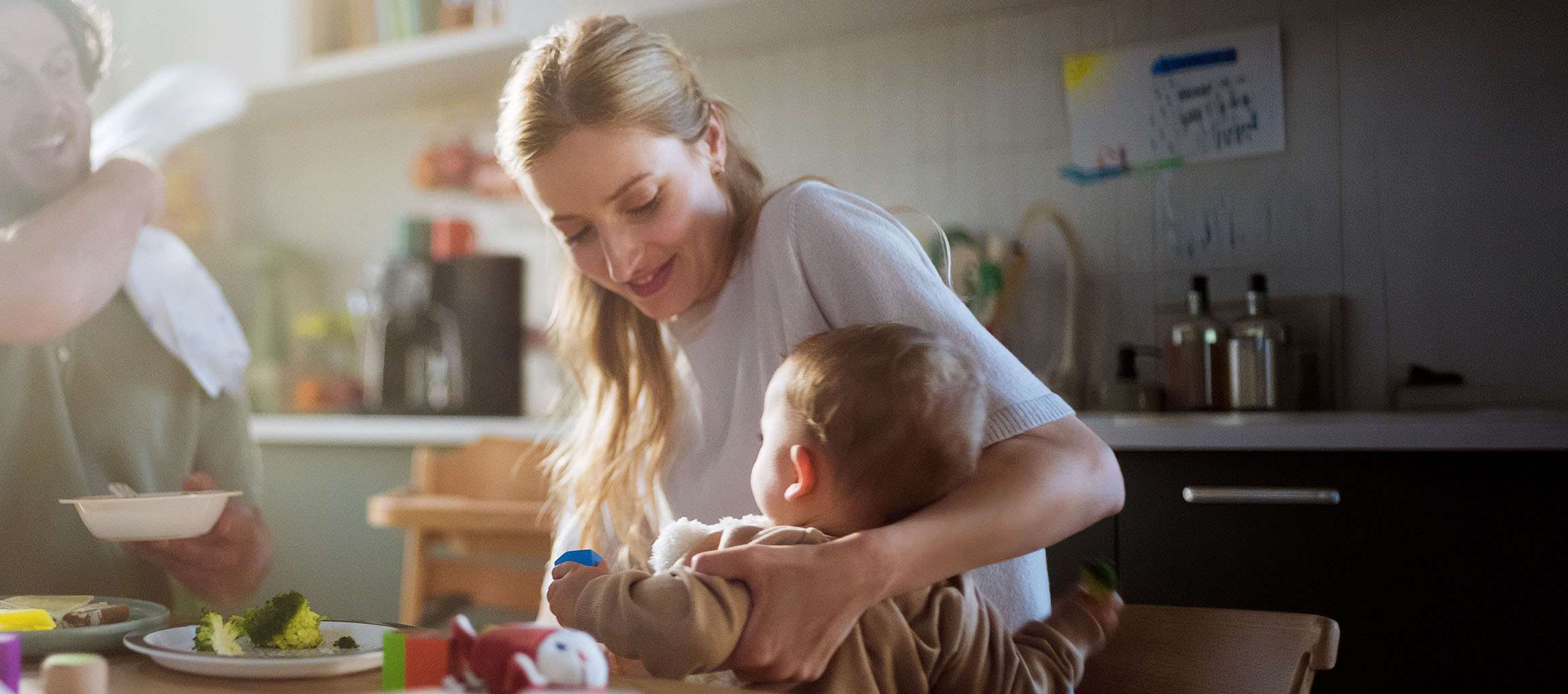 Family in kitchen, mother feeding baby, father eating broccoli from plate.
