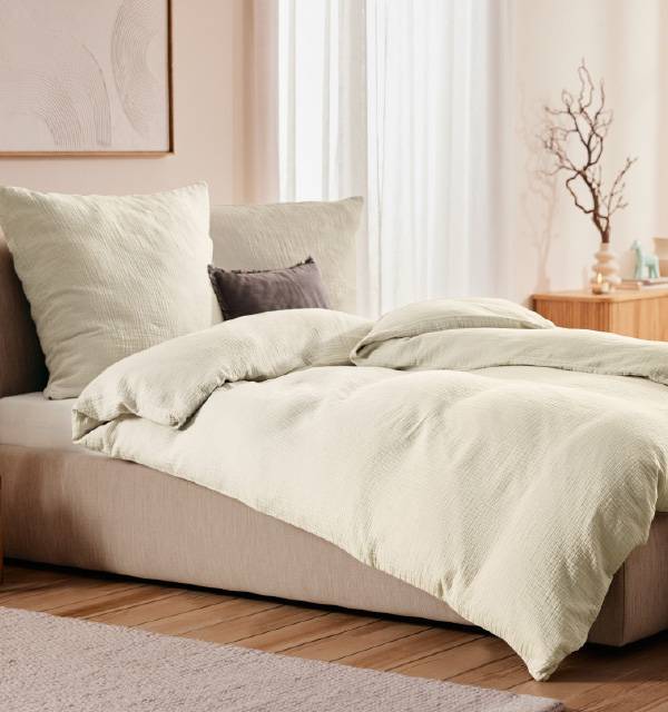 A cozy bedroom with a bed featuring light cream-colored bedding and pillows, with a wooden nightstand.