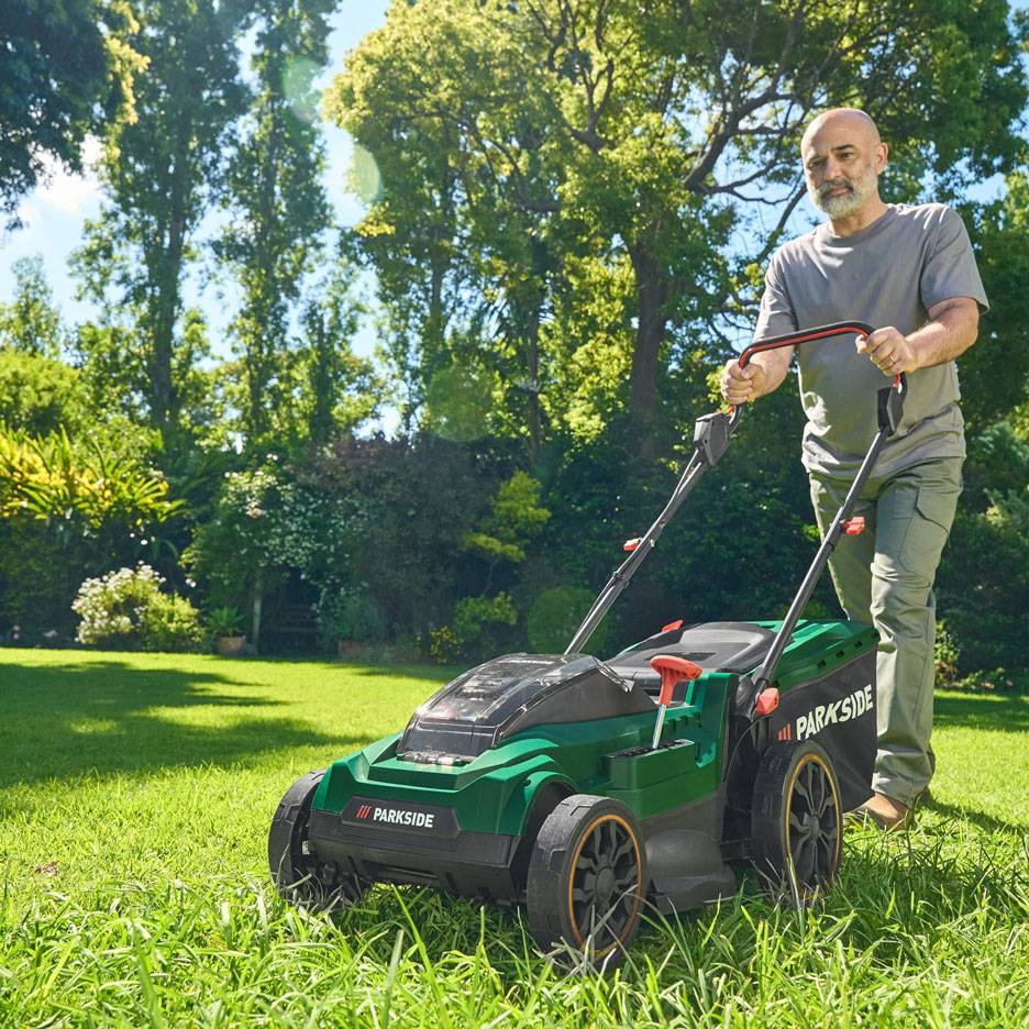 Man mowing lawn with a green lawnmower in a sunny garden.