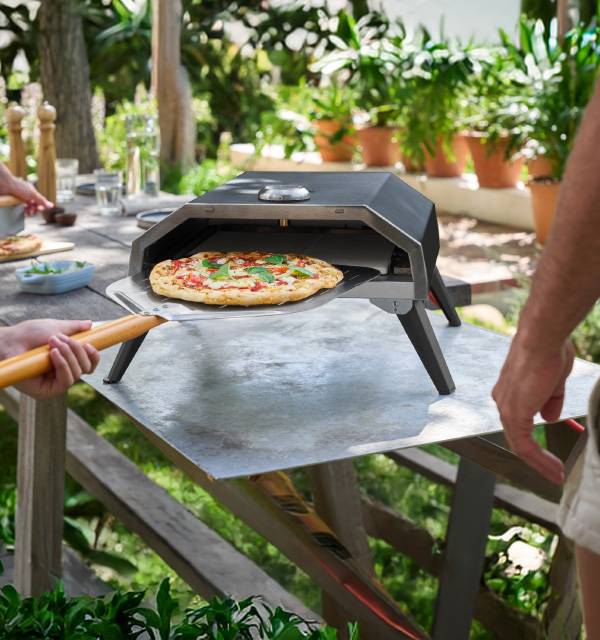 Outdoor pizza oven with a freshly baked pizza being removed with a peel in a garden setting.