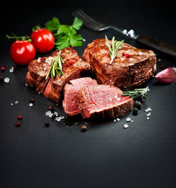 Cooked beef steaks with rosemary, tomatoes, garlic, and spices on a dark background.