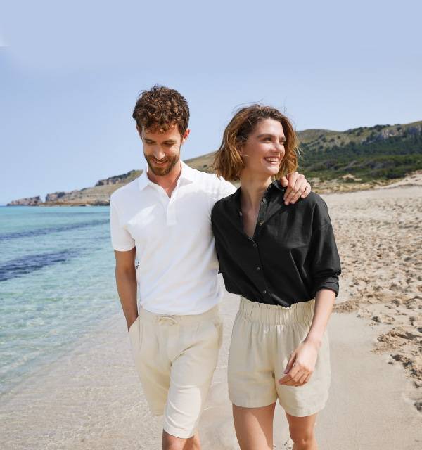 Couple in linen shorts and shirts walking on a sunny beach by the sea.