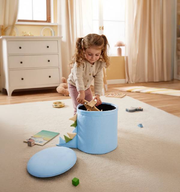 Little girl playing with a blue dinosaur toy storage box and wooden toys on a carpet.