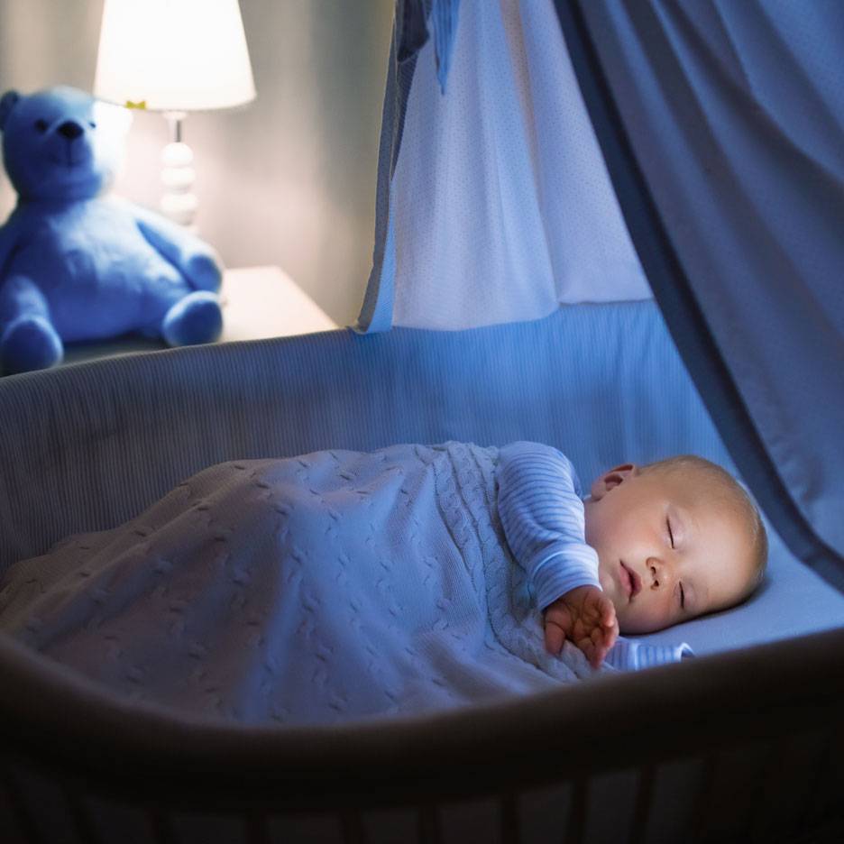 Sleeping baby in a crib with a blue teddy bear and a night light.