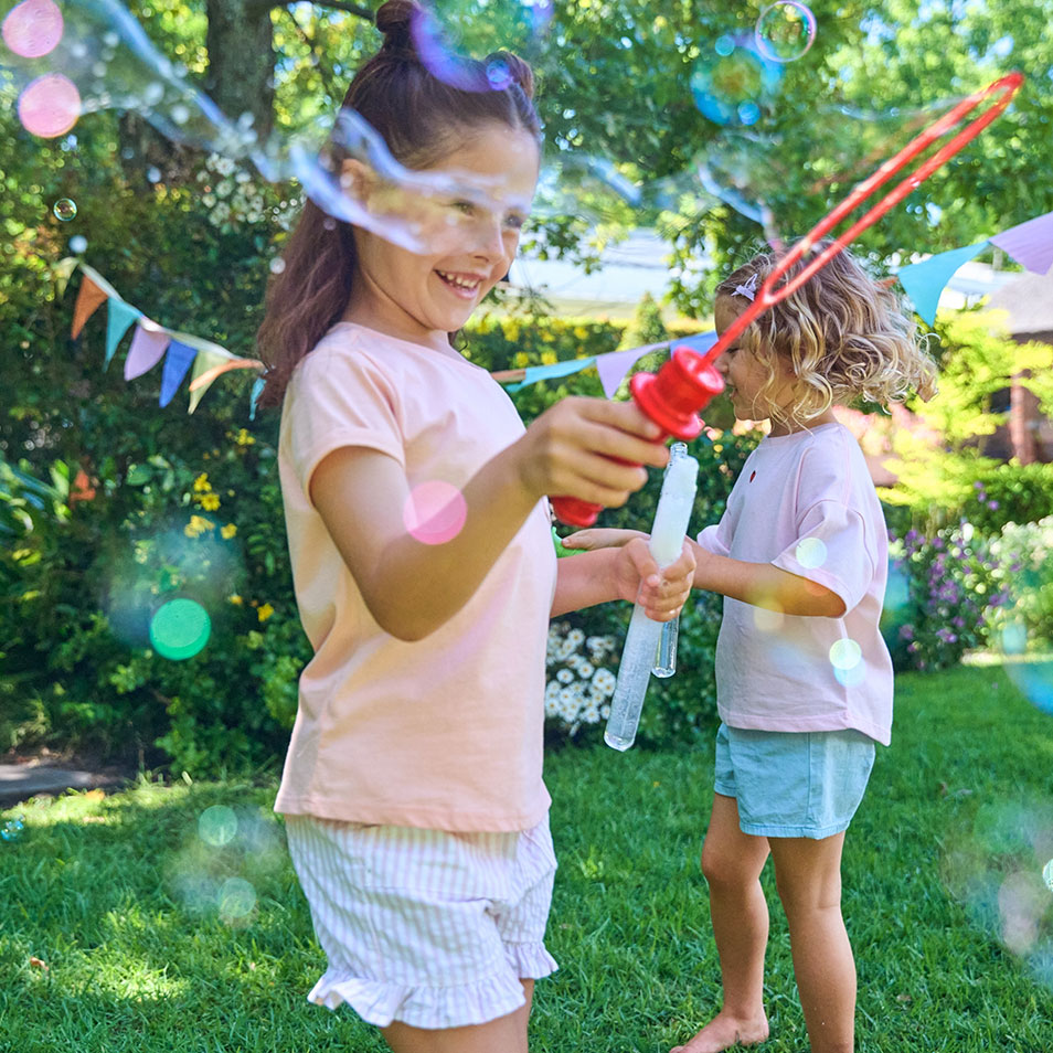 Two girls blowing bubbles in a garden, wearing summer clothes.