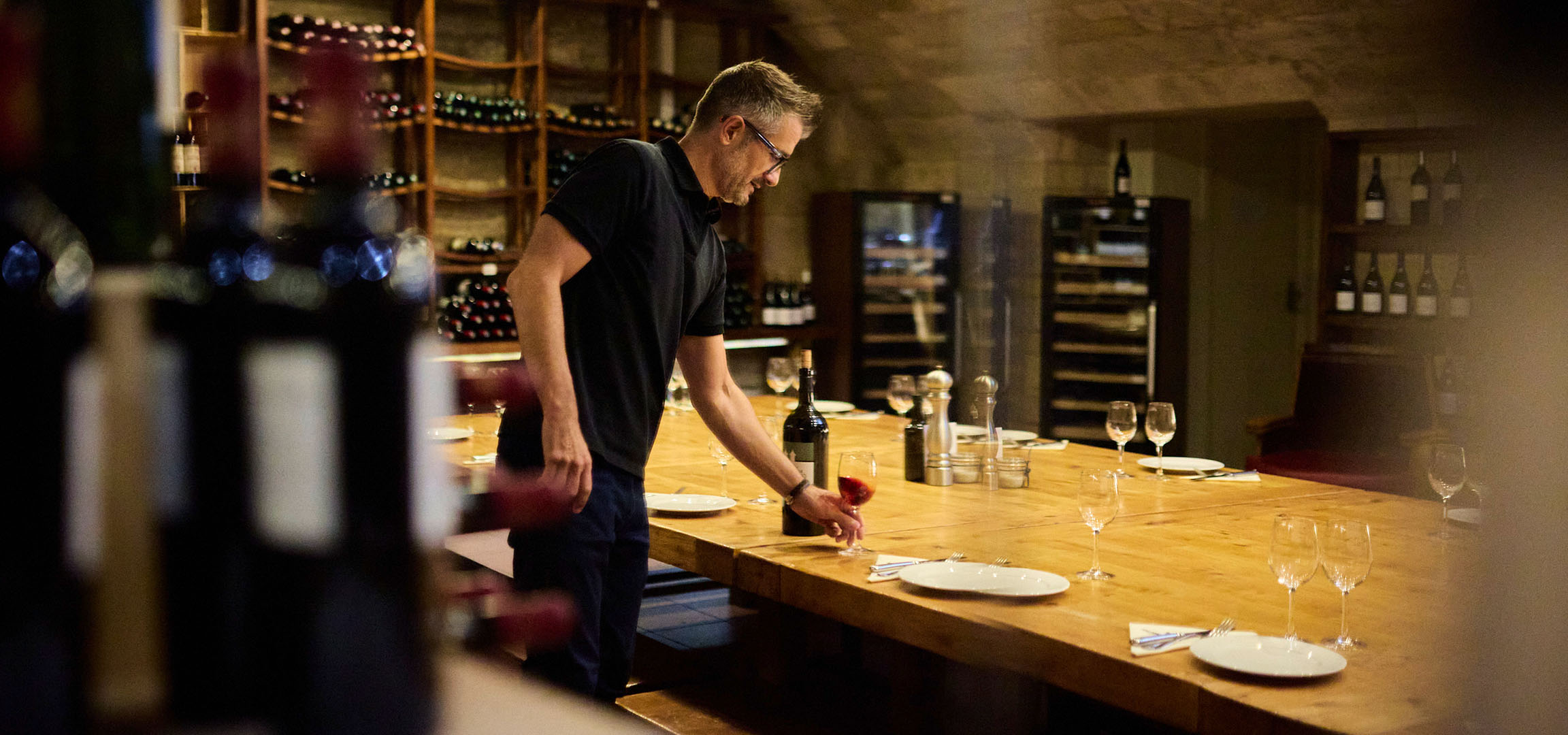 Man in a wine cellar, wine bottles on shelves and a table.