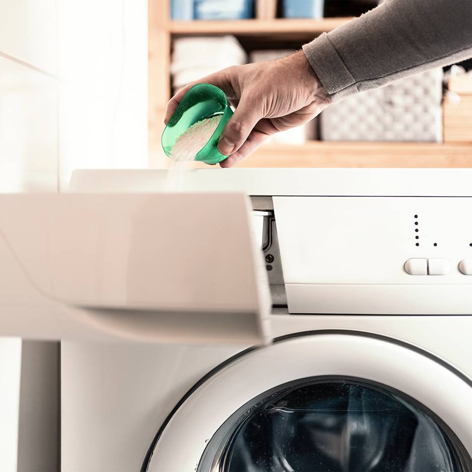 Hand with a scoop of laundry detergent over a washing machine.