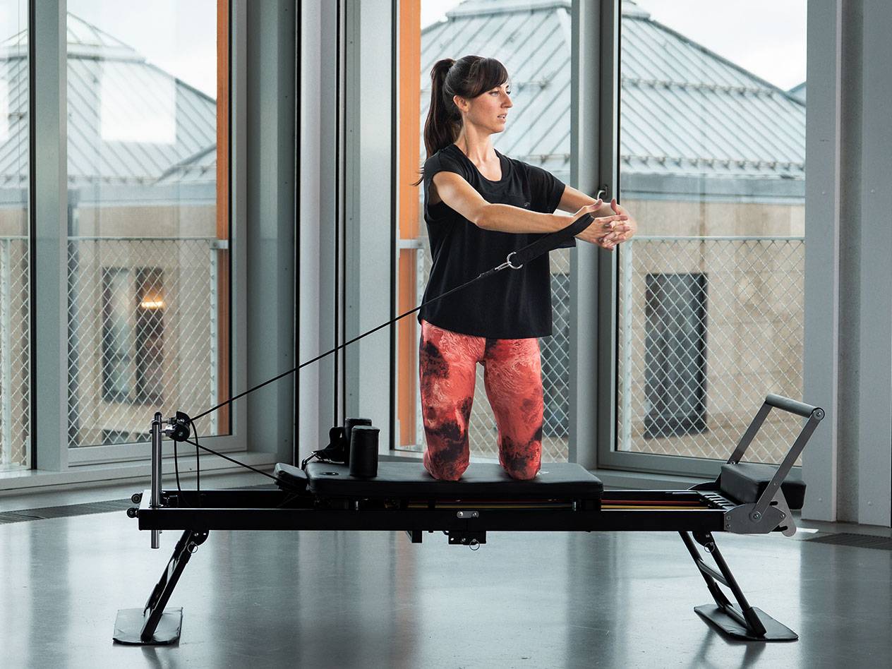 Woman doing Pilates on a black reformer machine in a bright room.