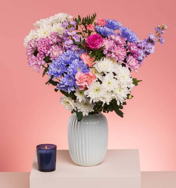 A vibrant bouquet of pink, purple, blue, and white flowers in a vase, with a lit candle.