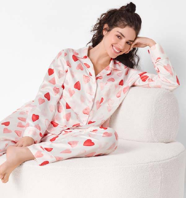 Woman in white and red heart-patterned satin pajamas, smiling and relaxing on a white chair.
