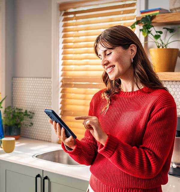 A smiling woman in a red sweater uses her smartphone in a modern kitchen.