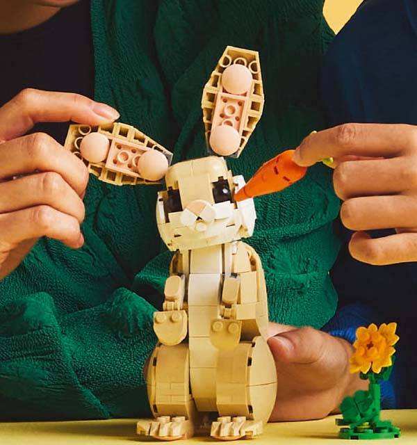 Hands assembling a toy brick bunny with ears and feeding it a carrot, with a flower nearby.