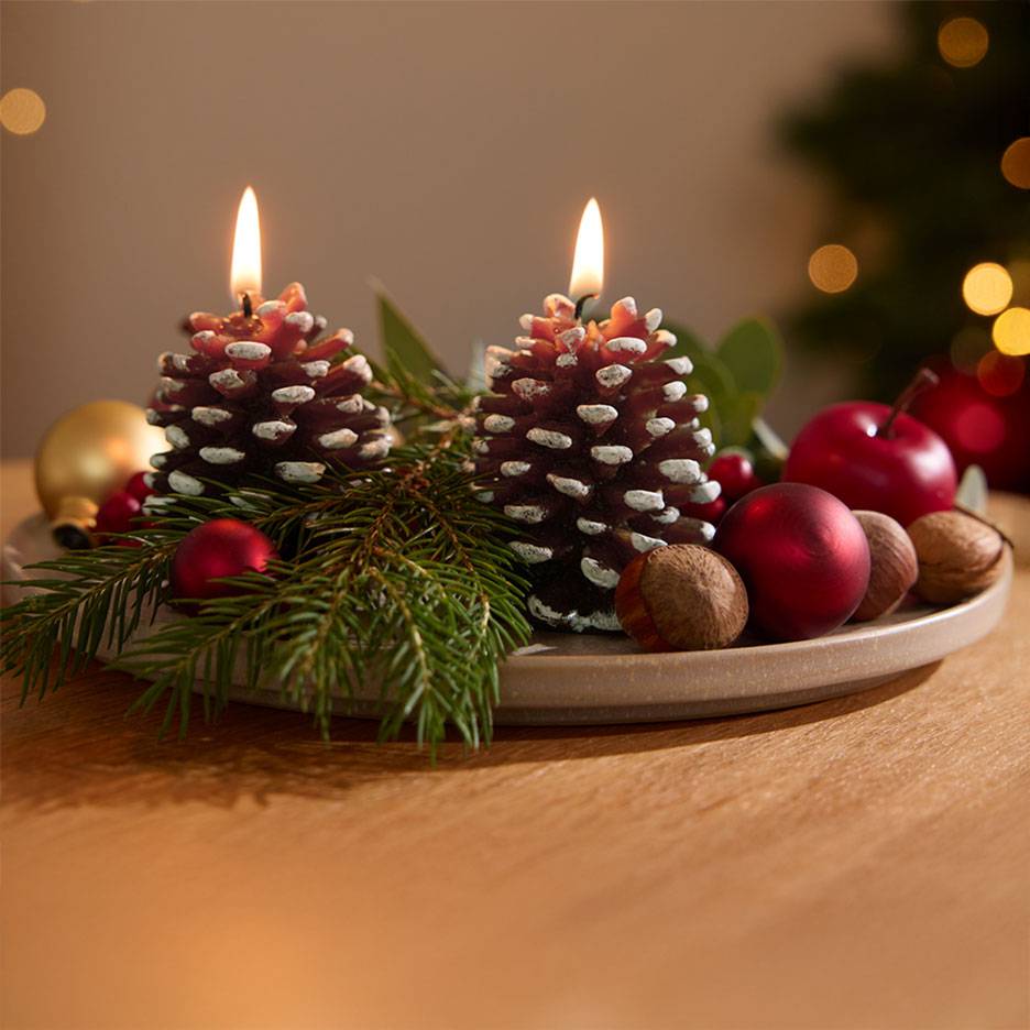 Advent decoration with two lit pine cone candles, fir branches, and Christmas ornaments on a plate.