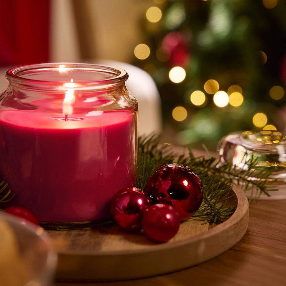 Red scented candle in a glass jar with Christmas baubles and fir branches on a wooden tray.
