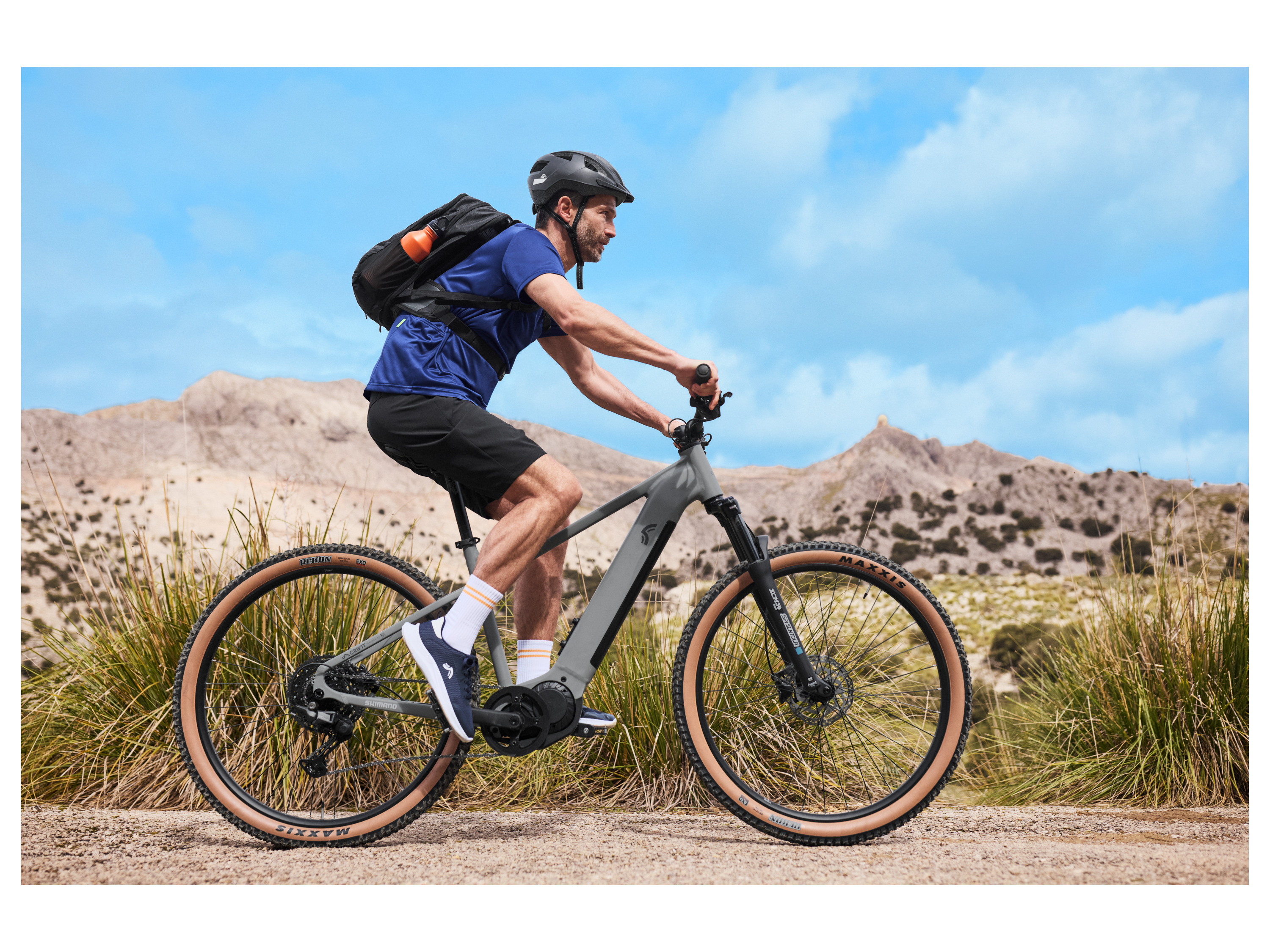 A man rides a gray Decathlon e-bike on a trail with tan-colored tires and a black backpack.