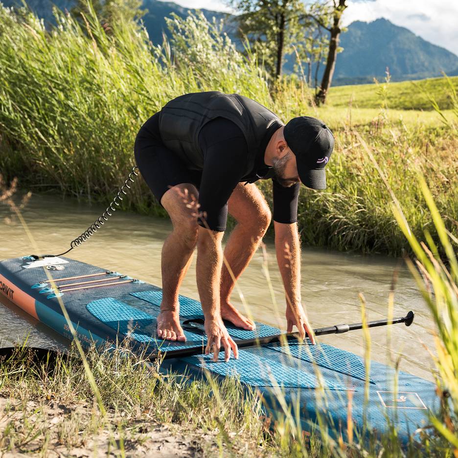 Man in wetsuit and life vest preparing for stand-up paddleboarding on a river.