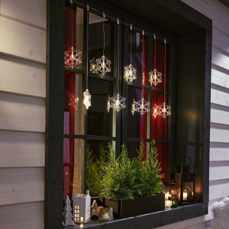 Snowflake string lights hanging in a window with small fir trees and candles on the sill.
