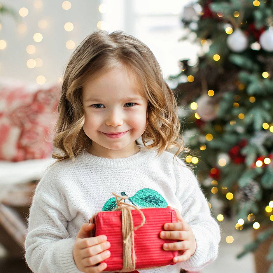 Smiling girl with a Christmas gift, with a lit Christmas tree in the background.