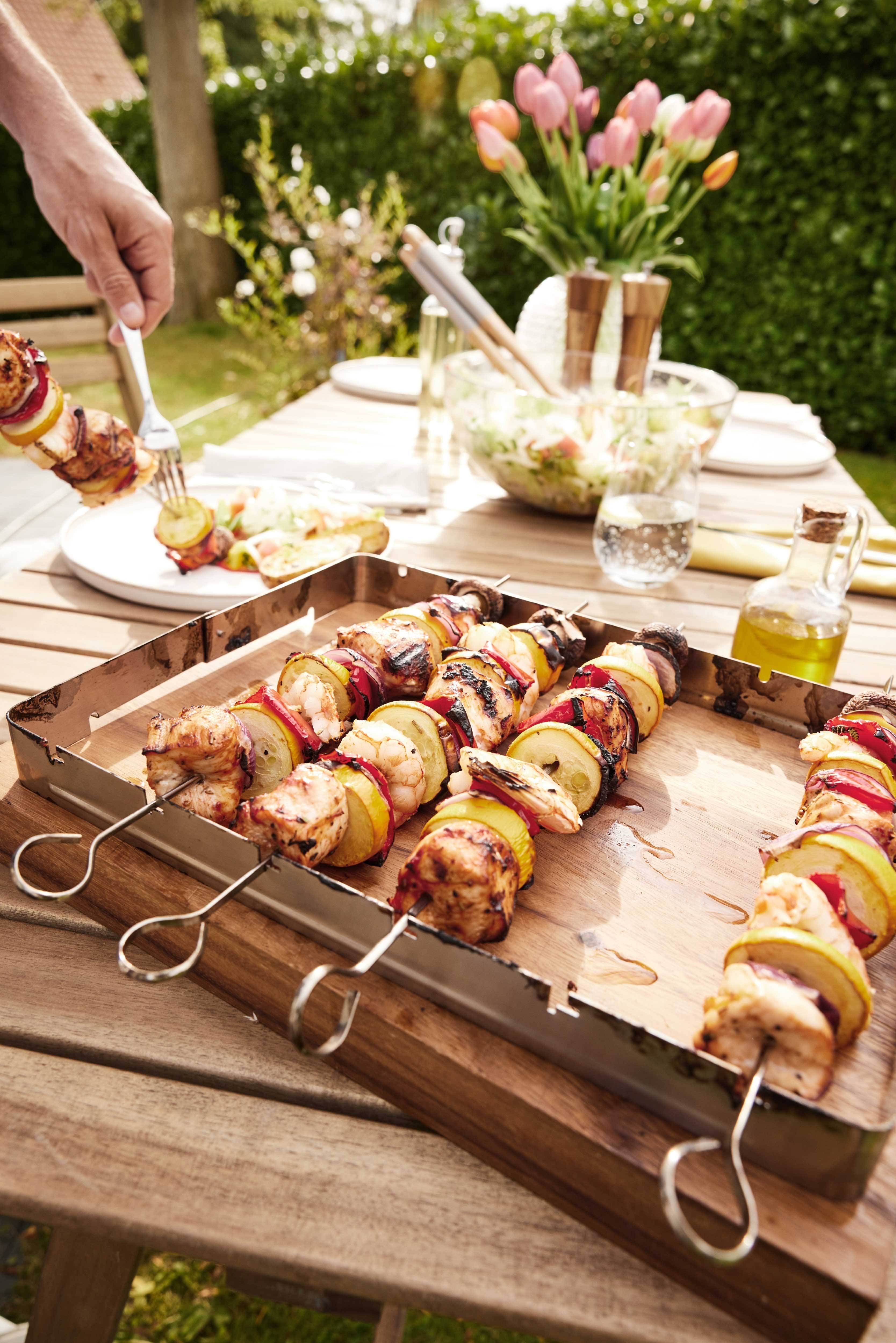 Chicken and vegetable skewers on an outdoor wooden table, with salad and tulips.