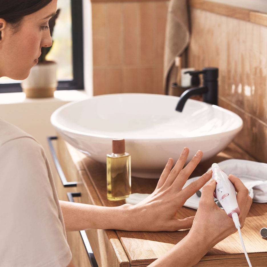 Woman using an electric manicure/pedicure set at a wooden vanity with a white sink.