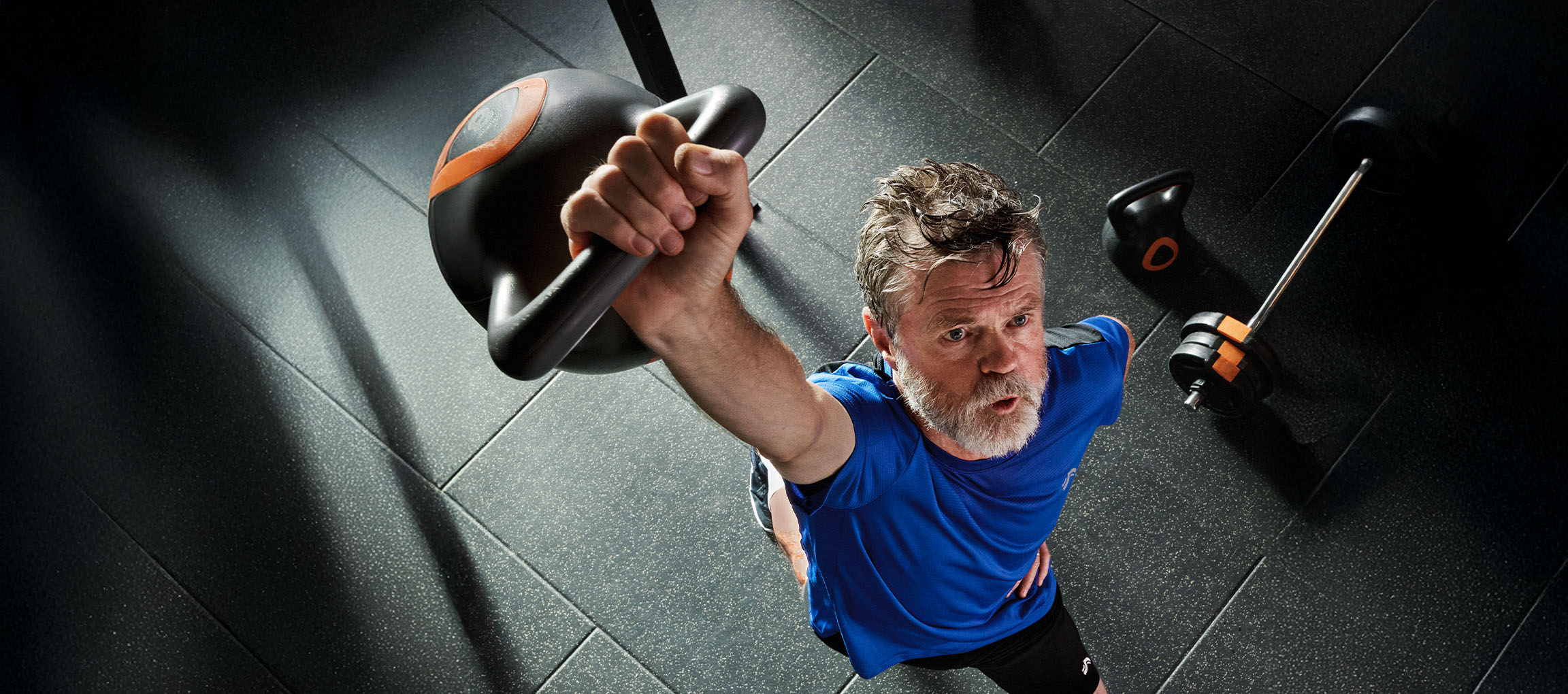 Man lifting a kettlebell in a gym, with a barbell and kettlebell on the floor.