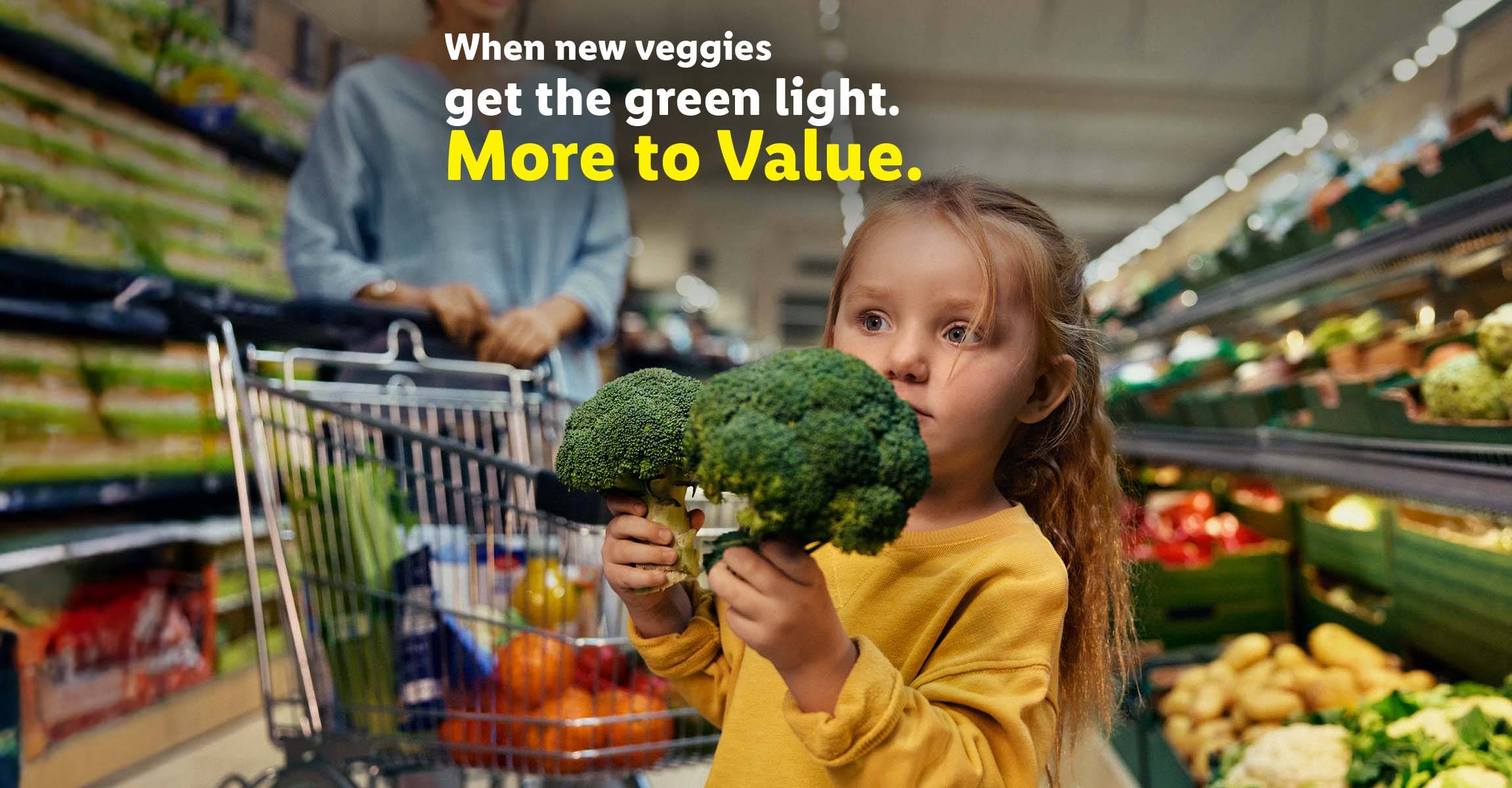 A child holding two broccoli heads in a grocery store with a shopping cart nearby.