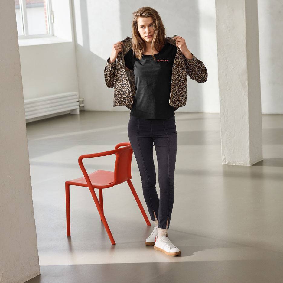 Woman in a black t-shirt with text, jeans, and a leopard print jacket, standing next to a red chair.
