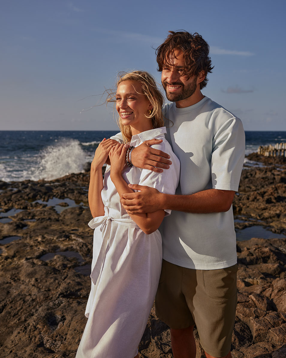 Couple on the beach, woman in white linen dress, man in light blue t-shirt and shorts.