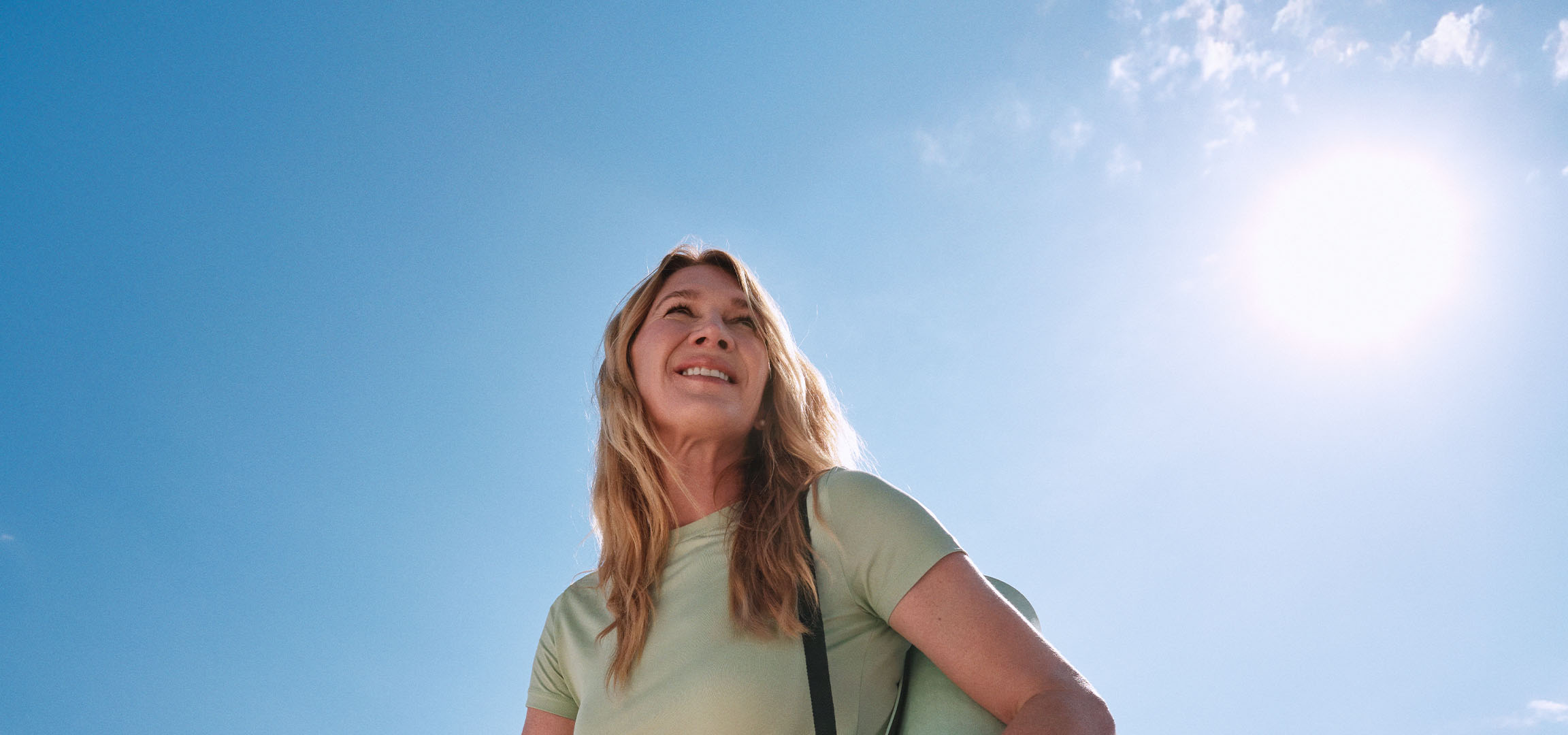Smiling blonde woman looking up at the sun under an open sky.