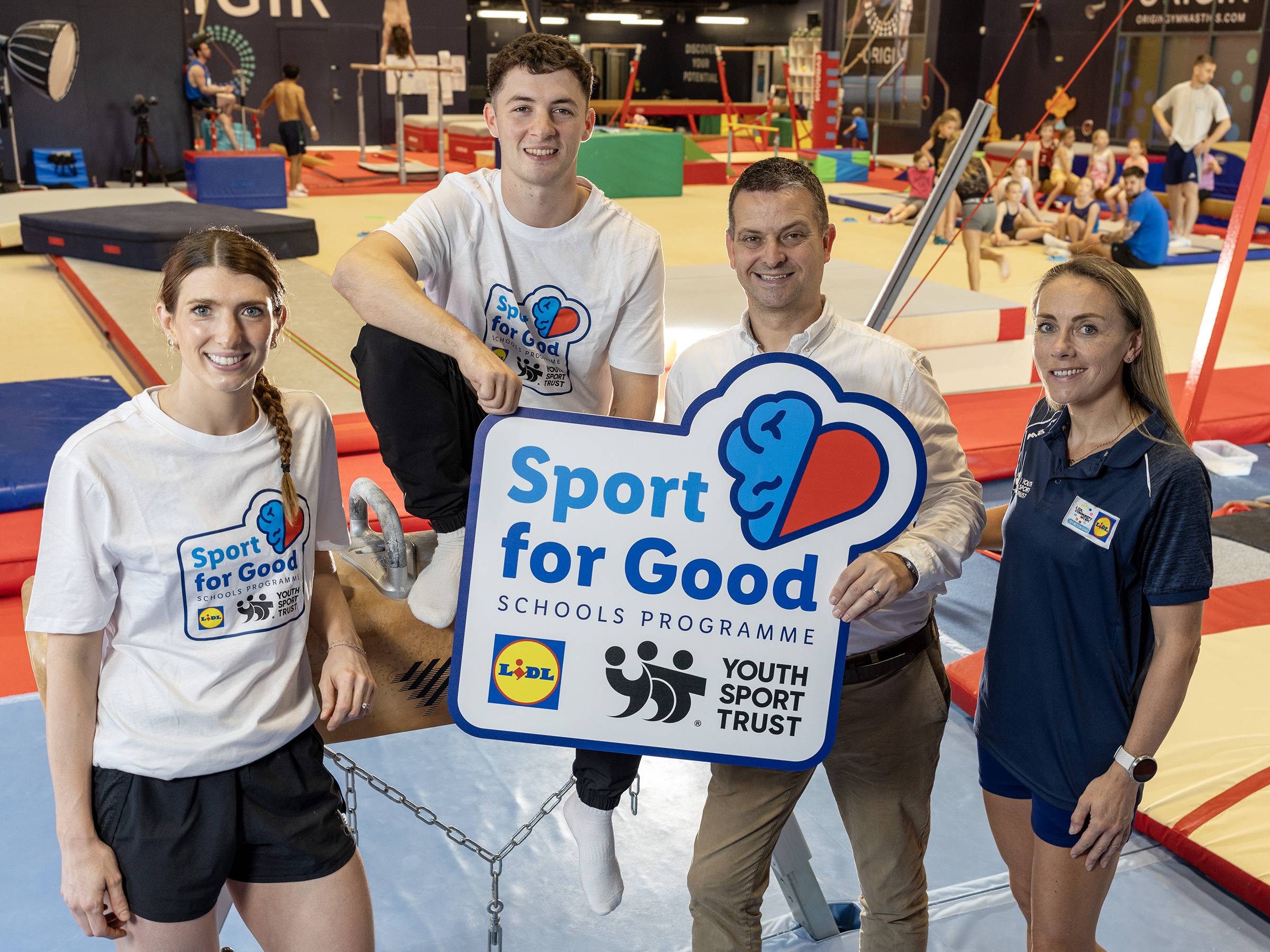 Four adults in a gym, two holding a 'Sport for Good' sign with Lidl and Youth Sport Trust logos.